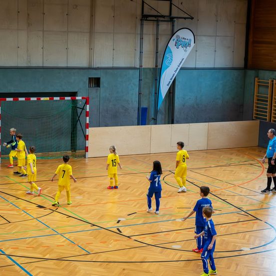 Eine Gruppe von Kindern mit nummerierten Trikots spielt Hallenfußball. Ein Kind steht in der Nähe des Torpfostens, während andere auf dem Feld positioniert sind.