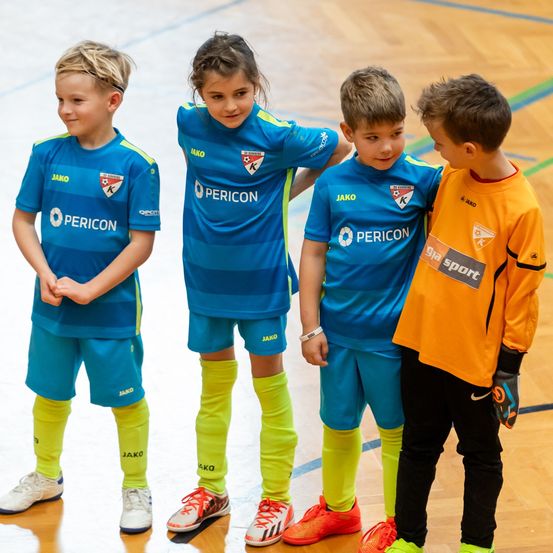 Vier Kinder stehen auf einem Spielfeld und tragen Sporttrikots. Drei tragen blaue Trikots mit 'Pericon' darauf, das vierte ein orangefarbenes Trikot mit 'ga sport'. Sie scheinen etwas zu besprechen.