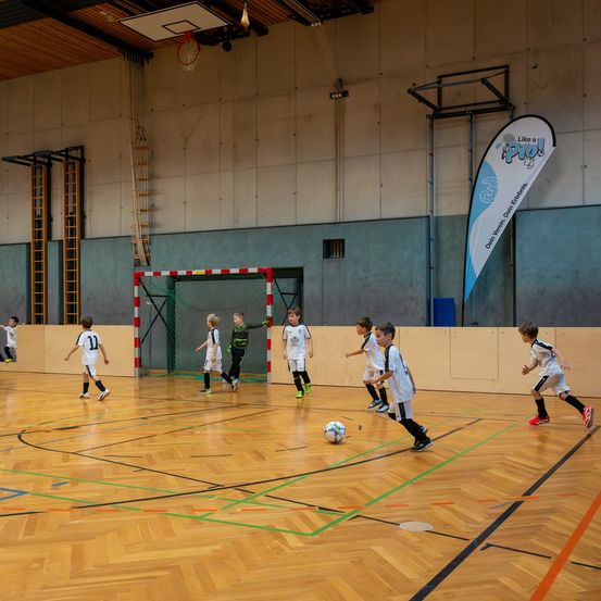 In einer Halle spielen eine Gruppe von Kindern in weißen Trikots Fußball. Ein Kind versucht, den Ball zu schießen. Hinter dem Tor steht ein Basketballkorb. Eine Fahne hängt an der Wand.