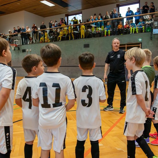 Ein Basketballtrainer spricht mit jungen Spielern in weißen Trikots mit den Nummern 3, 4, 11. Zuschauer beobachten vom Balkon.