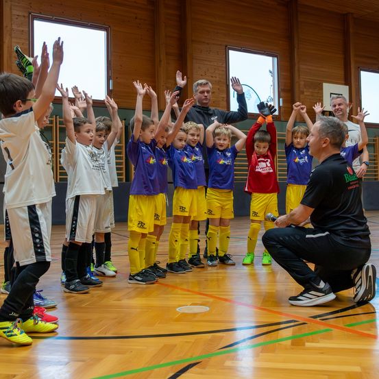 Eine Gruppe junger Jungen in Sportuniformen mit Trainern in einer Turnhalle. Sie heben ihre Hände, wahrscheinlich während einer Fußballtrainingseinheit.