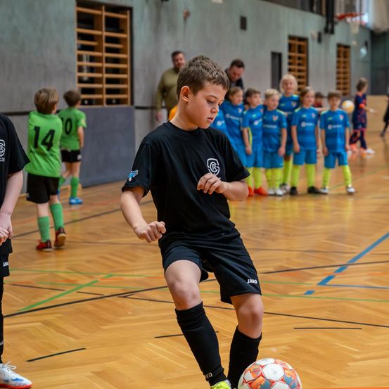 Ein Junge in einem schwarzen Trikot dribbelt einen Fußball auf einem Hof mit einem verschwommenen Hintergrund von Kindern in blauen Trikots.