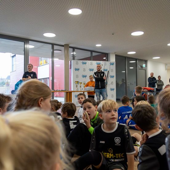 Eine Gruppe von Kindern ist drinnen versammelt und hört einem Mann zu, der hinter einem Tisch mit Trophäen steht. Ein anderer Mann steht in der Nähe einer Glaswand, und Menschen beobachten von den Seiten.