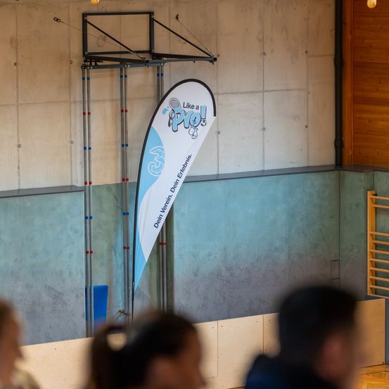 Ein Banner, das für einen Tennisclub wirbt, hängt an einer Wand in einer Sporthalle. Vier Menschen stehen davor, zwei Frauen und zwei Männer.