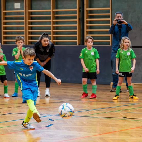 Mehrere Kinder spielen Fußball auf einem Hallenplatz, wobei ein Junge den Ball köpft. Zwei Kinder in grünen Trikots stehen dahinter, während eine Frau und ein Mann beobachten.