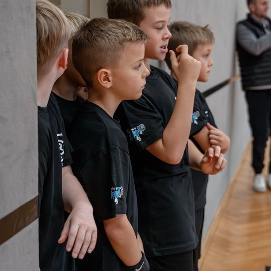 Eine Gruppe von Jungen in schwarzen T-Shirts steht in einer Reihe, möglicherweise während einer Teambesprechung. Sie schauen zur Seite, vielleicht zuhörend einem Redner.