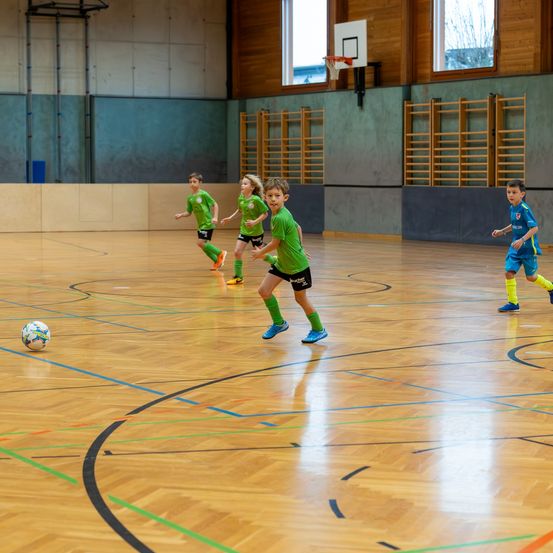 Fünf Kinder spielen Fußball auf einem Hallenplatz, drei in grünen Trikots und zwei in blauen Trikots. Ein Kind in Grün läuft auf den Ball zu.
