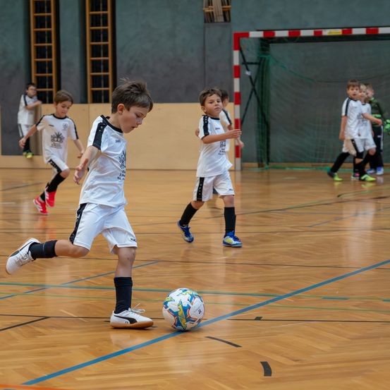Eine Gruppe von Kindern in weißen Fußballtrikots spielt auf einem Hallenplatz. Ein Kind tritt einen Ball mit seinem Fuß.