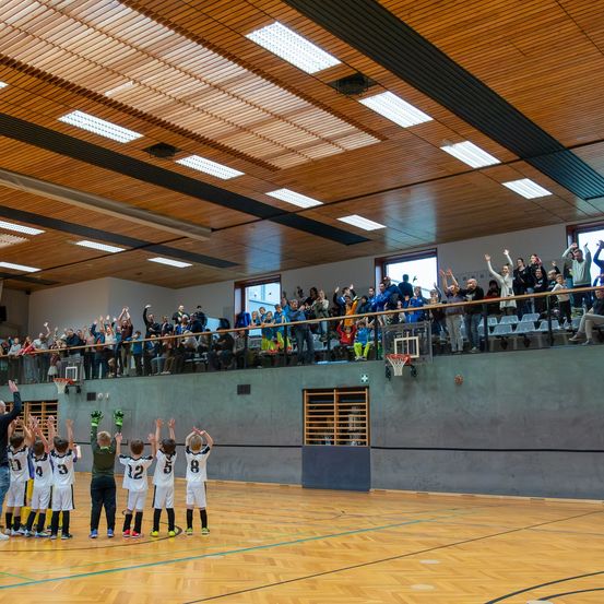 Eine Gruppe junger Basketballspieler feiert auf dem Spielfeld mit erhobenen Händen, während eine Menge auf den Tribünen zuschaut.