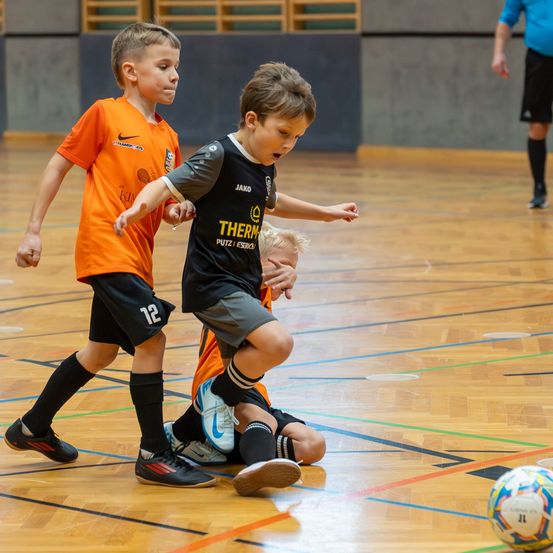 Drei Kinder spielen Hallenfußball. Ein Junge in Orange verfolgt einen anderen in Schwarz. Ein dritter Junge sitzt auf dem Boden, und ein Schiedsrichter beobachtet.