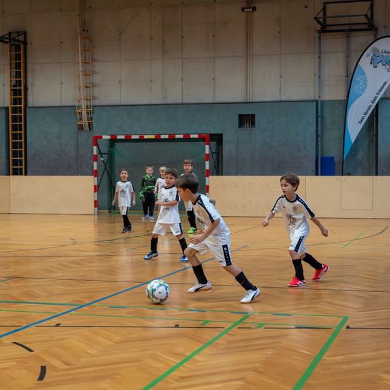 Eine Gruppe junger Jungen spielt Fußball auf einem Hallenplatz. Einige jagen dem Ball hinterher, während andere zuschauen. Der Platz hat Holzboden und blaue Markierungen.