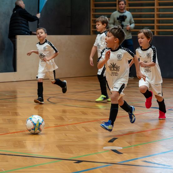 Vier kleine Jungen spielen Fußball auf einem Hallenplatz. Ein Junge läuft auf den Ball zu, während die anderen zuschauen. Ein Mann steht in der Nähe der Wand.