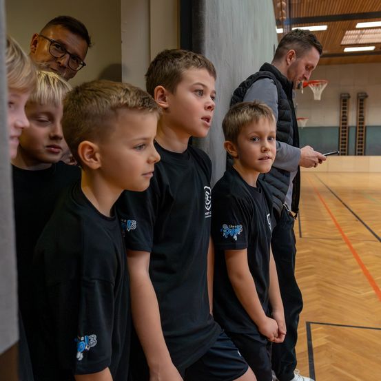 Eine Gruppe von Jungen in schwarzen T-Shirts steht in einer Reihe in einer Turnhalle und lauscht zwei Männern, von denen einer ein Handy hält. Die Turnhalle verfügt über ein Basketballfeld, Holzboden und Metallgeländer.