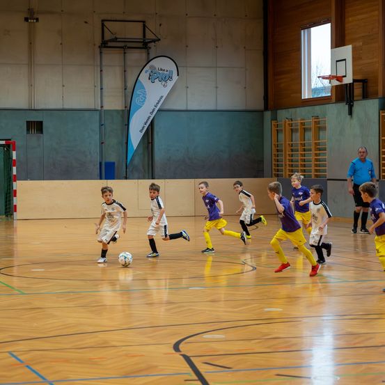 Mehrere junge Jungen in Sportuniformen spielen Hallenfußball. Ein Junge tritt den Ball, während andere darauf zulaufen. Ein Erwachsener beobachtet von der Seite.