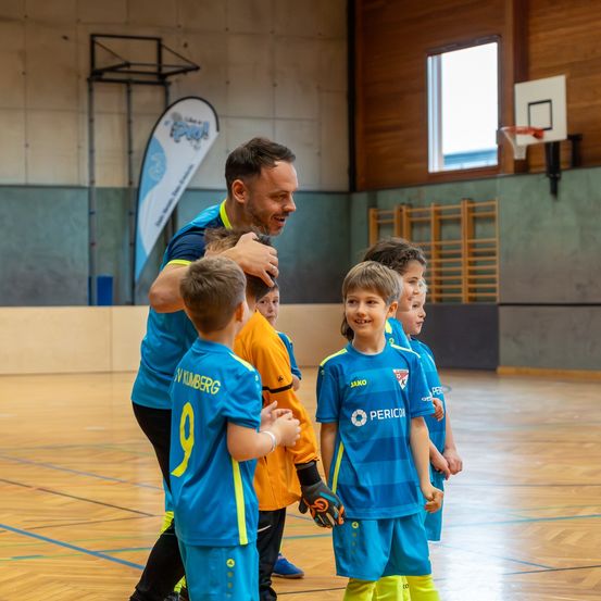 Ein Mann in einem blauen Shirt interagiert mit einer Gruppe junger Jungen in einer Sporthalle. Die Jungen tragen blaue Trikots mit Nummern und gelben Akzenten.