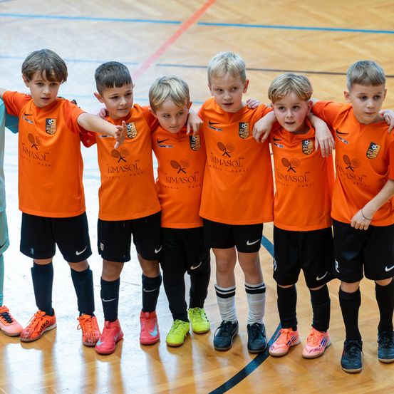 Eine Gruppe junger Jungen in orangefarbenen Trikots mit dem Text 'KIMASOL' steht zusammen auf einem Platz. Sie sind wahrscheinlich eine Fußballmannschaft und tragen schwarze Shorts und Socken. Ihre Schuhe sind farbenfroh.