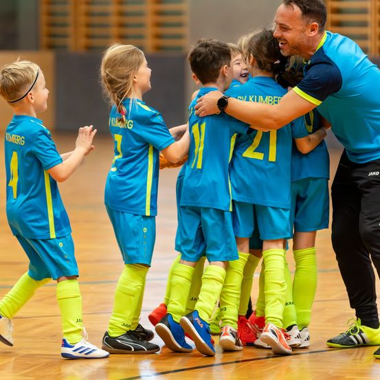 Eine Gruppe von Kindern in blauen und gelben Uniformen feiert auf einem Basketballplatz. Ein Trainer umarmt ein Mädchen.