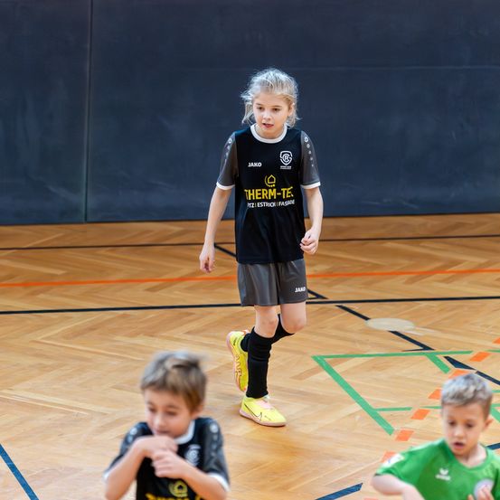 Ein Mädchen in einem schwarzen und gelben Fußballtrikot mit Text läuft auf einem Spielfeld, während zwei Jungen zusehen.