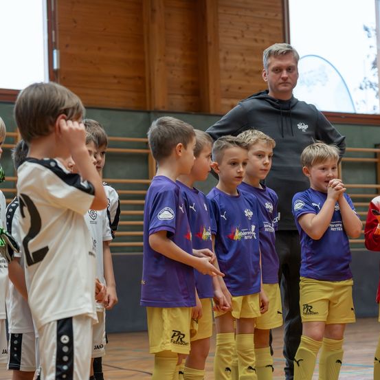 Eine Gruppe junger Jungen in Fußballtrikots, einige in Purpur und Gelb, andere in Weiß, stehen in einer Reihe in einer Turnhalle, mit einem Mann in einer schwarzen Kapuzenjacke hinter ihnen.