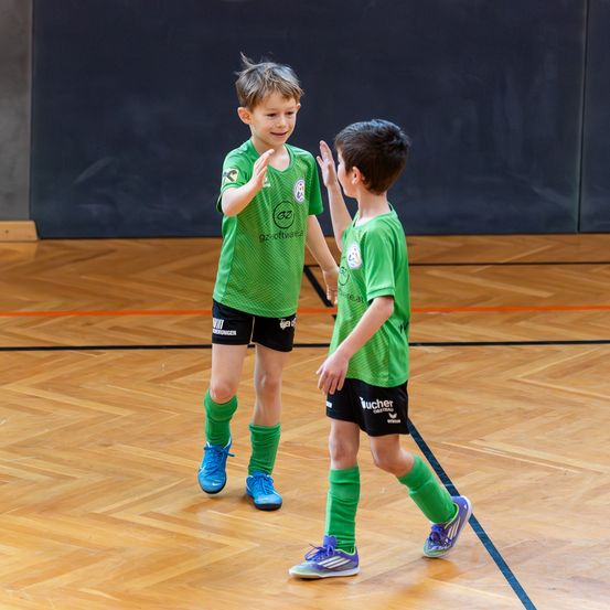 Zwei junge Jungen in grünen Fußballtrikots stehen auf einem Holzboden, lächeln und geben sich High-Fives.