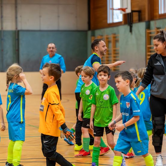 Kinder in Sporttrikots, wahrscheinlich Fußball, versammeln sich in einer Turnhalle. Zwei Frauen und zwei Männer stehen vor ihnen. Eine Basketballkorb ist an der Wand montiert.