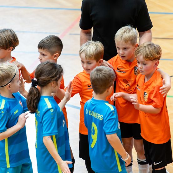Eine Gruppe junger Kinder in Sporttrikots steht in einer Turnhalle, bereit für ein Spiel. Einige tragen orangefarbene Trikots, andere blaue. Ein Trainer steht hinter ihnen.