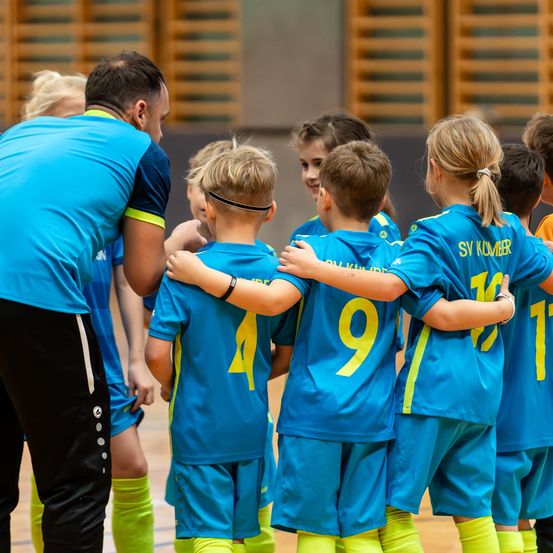 Eine Gruppe junger Kinder in blauen und gelben Sporttrikots schmiedet sich zusammen, ein Trainer ist in der Nähe. Die Kinder haben Nummern auf ihren Trikots, die von 1 bis 17 reichen. Sie scheinen sich auf einem Sportplatz zu befinden.