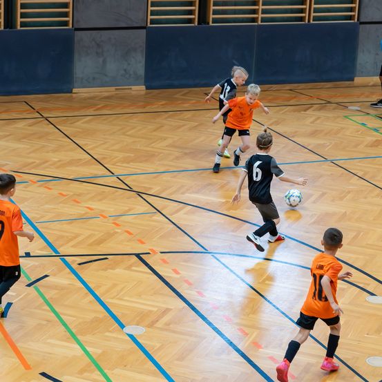 Eine Gruppe junger Jungen spielt Fußball auf einem Hallenplatz. Sie tragen orangefarbene und schwarze Trikots mit Nummern. Ein Junge tritt den Ball.