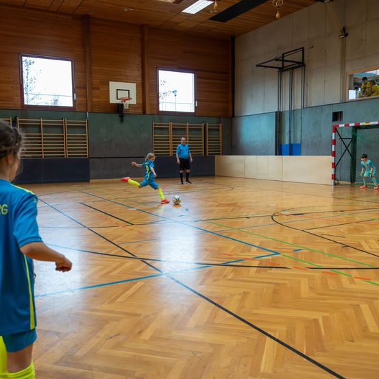 In einer Indoor-Sporthalle spielen mehrere Kinder Fußball. Ein Kind tritt den Ball. Die Halle hat Holzböden und Wände.