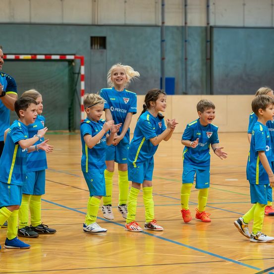 Mehrere Kinder in blauen und gelben Uniformen stehen auf einem Basketballplatz, lächeln und schauen in die Kamera. Ein Mann beobachtet sie von hinten.