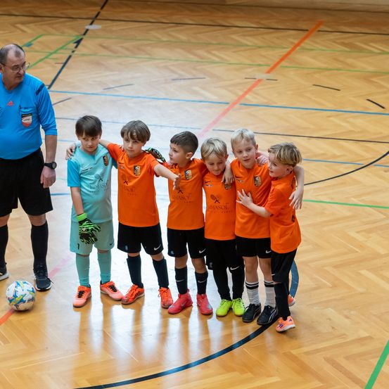 Eine Gruppe junger Fußballspieler in orangefarbenen Trikots steht auf einem Spielfeld zusammen, ein Mann in einem blauen Hemd steht hinter ihnen.