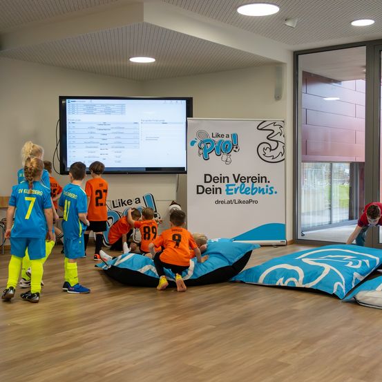 Kinder in Sporttrikots beobachten einen Monitor, während einige auf Bohnensäcken sitzen. Ein Plakat im Hintergrund sagt 'Like a Pro'.