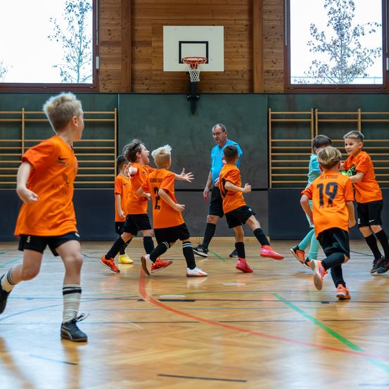 Kinder in orange Trikots spielen Hallenfußball. Ein Mann in einem blauen Hemd beobachtet sie. Der Basketballkorb ist an der Wand montiert.