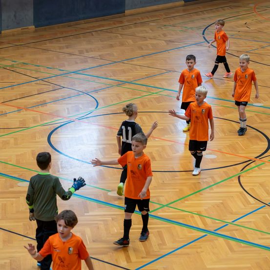 Eine Gruppe junger Kinder in orangefarbenen Trikots spielt Fußball auf einem Hallenplatz. Ein Kind hält einen Fußball, andere sind in verschiedenen Positionen.