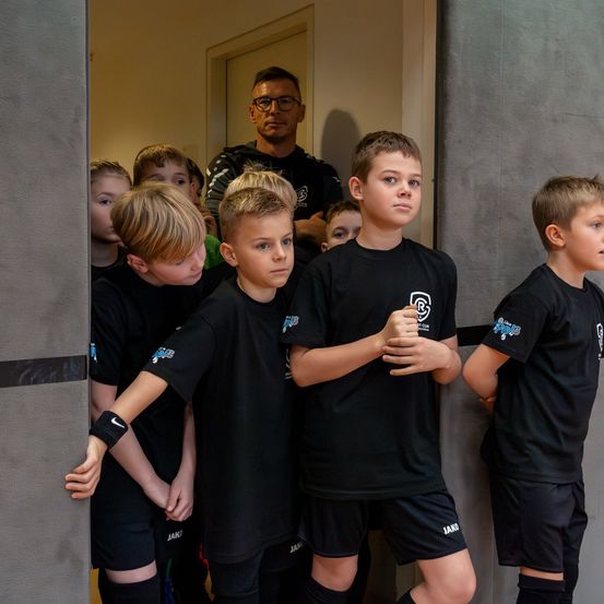 Eine Gruppe von Jungen in schwarzen Trikots steht zusammen in einem Raum und blickt seitwärts. Hinter ihnen steht ein Mann mit Brille in der Nähe der Tür und beobachtet.
