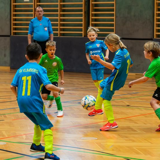 Mehrere Kinder in Sportuniformen spielen auf einem Spielfeld Fußball. Ein Junge tritt den Ball, während ein Mädchen versucht, ihn zu blockieren. Ein Schiedsrichter beobachtet von hinten.