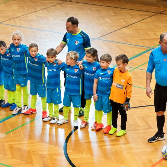 Eine Gruppe junger Kinder, die blaue und gelbe Sporttrikots tragen, steht in einer Reihe mit einem Trainer auf einem Platz. Ein Fußball liegt in der Nähe.