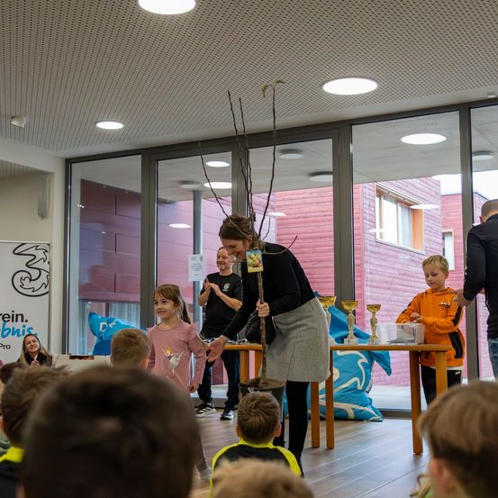 Eine Frau hält einen Stock, während ein Mädchen vor einer Gruppe von Kindern näher kommt. Dahinter steht ein Mann in der Nähe eines Tisches mit Trophäen.