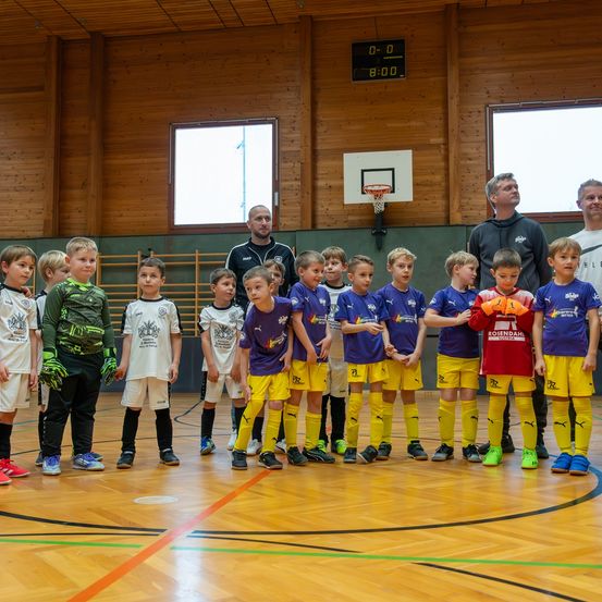 Eine Gruppe von Kindern und Erwachsenen steht auf einem Basketballplatz, trägt Sportuniformen und Turnschuhe. Sie scheinen auf etwas zu warten. Der Platz hat einen Holzboden, und es gibt Fenster und ein Anzeigebrett.