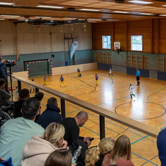 Ein Hallenfußballspiel ist im Gange mit jungen Spielern. Zuschauer beobachten von den Seitenlinien, und ein Fotograf hält den Moment fest. Der Platz hat blaue und orange Markierungen.