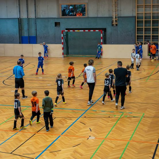 Eine Gruppe junger Kinder spielt ein Spiel Fußball auf einem Hallenplatz. Sie tragen Trikots mit Nummern und Turnschuhe. Sie stehen vor einem Netz, ein Trainer steht in der Nähe. An der Wand befindet sich ein Bilderrahmen, und rechts steht eine Leiter.