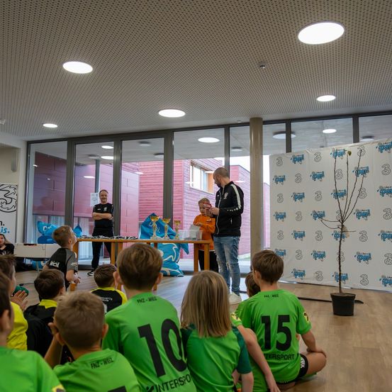 Eine Gruppe von Kindern in grünen Trikots sitzt in einem Raum und hört einem Mann zu. Ein Tisch mit Trophäen steht hinter ihnen. Ein weiterer Mann steht in der Nähe. Eine Tafel mit Text und einer Pflanze befindet sich rechts.