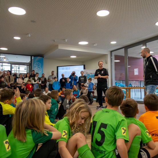 Mehrere junge Kinder sitzen vor einem Mann, der eine Rede hält, auf dem Boden. Dahinter stehen noch viele weitere Kinder und Erwachsene.