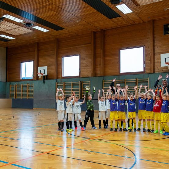 Eine Gruppe junger Kinder in Sportuniformen, wahrscheinlich Fußballspieler, heben ihre Hände und lächeln in einer Turnhalle. Der Boden ist mit Linien markiert.