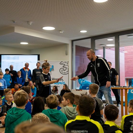 Ein Mann überreicht einem jungen Jungen eine Karte während einer Veranstaltung in einem Raum mit Glaswänden. Der Junge hält eine Karte und schaut den Mann an. Hinter ihnen beobachten eine Gruppe von Kindern und Erwachsenen. An der Wand befindet sich eine Projektionsleinwand und ein Banner mit einem Logo.