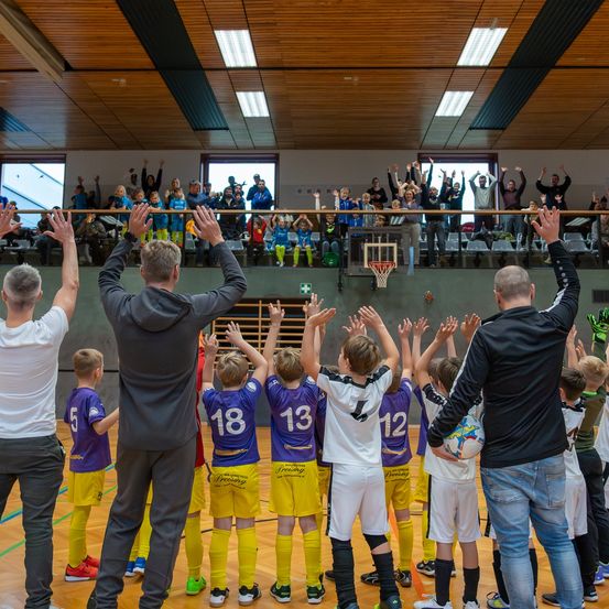 Eine Gruppe von Kindern in Sporttrikots steht auf einem Basketballplatz und hebt die Hände. Zwei Erwachsene vor ihnen heben ebenfalls die Hände. Ein Publikum beobachtet von den Tribünen.