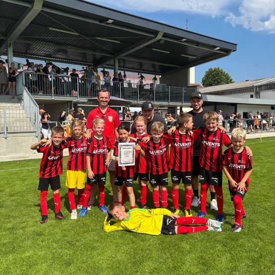 Eine Fußballmannschaft posiert mit ihrem Trainer und einem Zertifikat auf einem Feld, mit Zuschauern im Hintergrund.