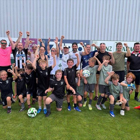 Eine Gruppe von Fußballspielern, Erwachsene und Kinder, posiert für ein Foto mit erhobenen Armen und Fäusten auf einem Rasenfeld.