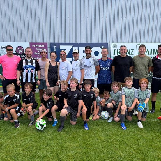 Eine Gruppe von Menschen, darunter junge Jungen und Männer, posiert für ein Foto auf einem Fußballplatz. Sie tragen Trikots mit verschiedenen Team-Logos und Nummern.