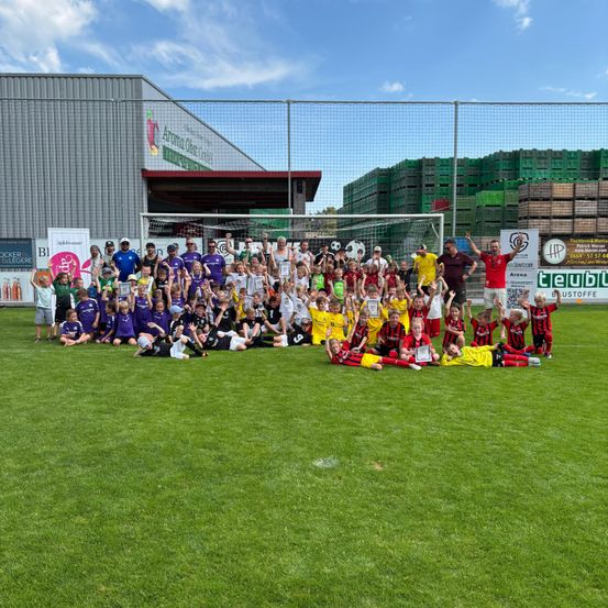 Eine große Gruppe von Kindern und Erwachsenen, gekleidet in Sportkleidung, posiert für ein Foto auf einem Fußballplatz. Hinter ihnen ein Gebäude mit einem Schild. In der Ferne ein Zaun mit Werbung.